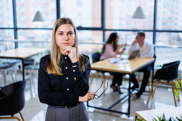 Молодая женщина в черной рубашке с яркими эмоциями на лице, она стоит на фоне своих коллег и жестикулирует руками - Фото, изображение