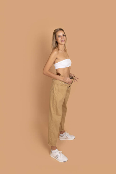Young blonde woman in good shape smiling wearing big oversize pants to show weight loss on skin colored background. Keeping a healthy diet, exercising - Photo, Image