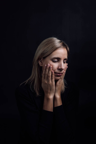 Pretty depressed woman with hands on cheeks in black dress on black background. Victim of physical and psychological abuse. Gaslighting. Relative aggression - Photo, image