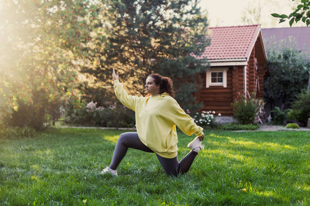 Young woman wearing sports clothes doing yoga exercises on backyard on green grass with wooden country house and high trees in background. Body positive - Foto, immagini