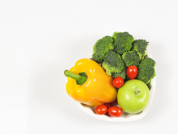 Bovenaanzicht of platte lay van groenten capsicum, broccoli en tomaten en appel in hart vorm plaat op witte achtergrond met kopieerruimte, gezonde levensstijl en gewichtsverlies concept. - Foto, afbeelding
