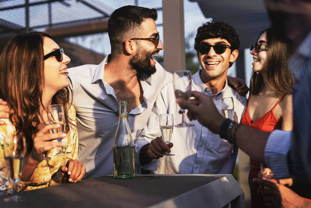 Najlepsi przyjaciele zbierający się na plenerowej imprezie beztroski zachód słońca razem rozmawiać, uśmiechając się i żartując razem pijąc musującego szampana - millenials grupa beztroska w weekend stojąc na stole bar - Zdjęcie, obraz