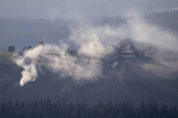 Hegyi táj. Sok füst jön ki egy kéményből egy régi házból Lupcinából, Romániából. - Fotó, kép