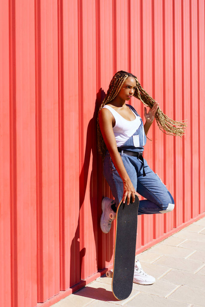 黒の女性はカジュアルな服装,赤い都市の壁の背景にスケートボードと. - 写真・画像