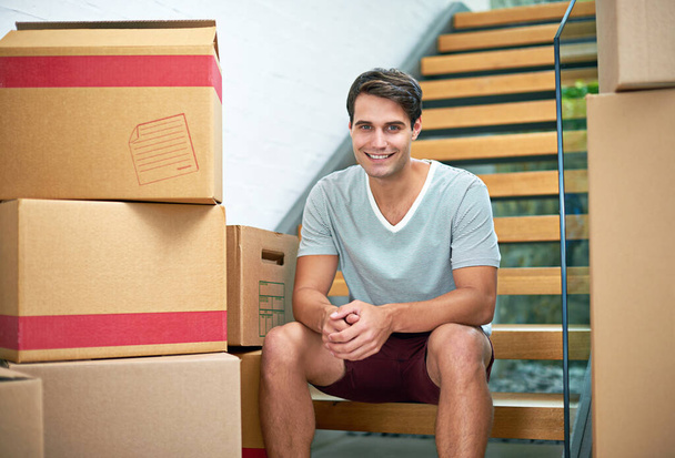 Rozpoczęcie nowego rozdziału. Portret młodego mężczyzny siedzącego w nowym domu otoczonego kartonowymi pudełkami. - Zdjęcie, obraz