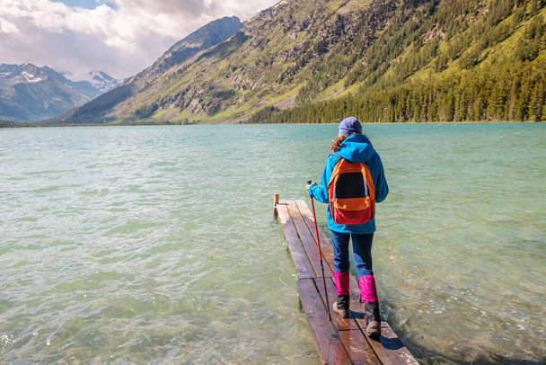 ハイカーの女の子旅行者は、風光明媚な山の湖で漁船のための木製の桟橋に立っています - 写真・画像