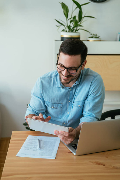 Веселый улыбающийся бизнесмен смотрит или читает что-то на своем цифровом планшете, сидя за столом с ноутбуком и документами и работая дома - Фото, изображение