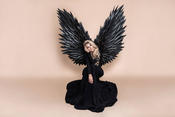 Elegant fashion. Stunning blonde woman in elegant long black dress and big wings on beige background in studio. Luxury evening fashion. Glamour fashion model. Classy woman. Woman with wings. Angel. - Photo, Image