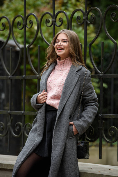Μοντέρνο και όμορφο χαμογελαστό νεαρή γυναίκα 16-20 ετών, ντυμένη με ένα ροζ πλεκτό πουλόβερ, γυαλιά, σκούρα φούστα και μια γκρι κλασική καμπαρντίνα. - Φωτογραφία, εικόνα