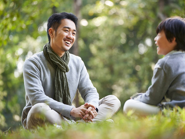 азіатський батько і син, сидячи на траві, приємно розмовляють на відкритому повітрі в парку
 - Фото, зображення