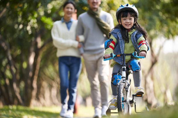 Kasklı ve korunaklı küçük Asyalı kız şehir parkında bisiklet sürüyor ve ailesi arkadan izliyor. - Fotoğraf, Görsel