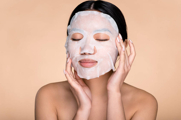 Sensual photo of beautiful young asian woman with with eyes closed, applying cotton facial moisturizing mask on face, takes care of skin, prevents wrinkles, stands on isolated beige background - Foto, imagen