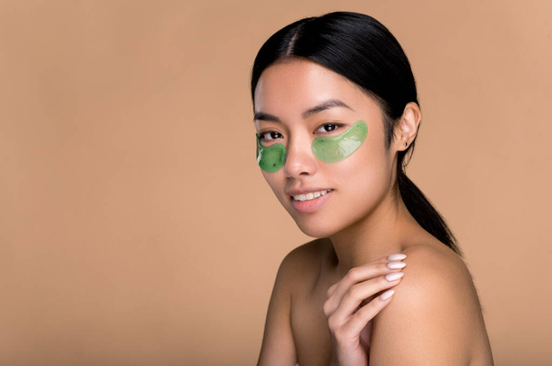 アンチエイジングの美容方法。完璧な手入れの行き届いた肌を持つ中国の女性は、目の下に化粧品コラーゲン緑のパッチを適用し、目の周りの皮膚をケアし、しわを防ぎ、ベージュの背景を隔離し、笑顔 - 写真・画像