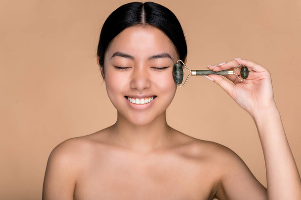 Asian beautiful girl holding a jade massage roller for facial skin in her hand, makes anti-aging measures, wrinkle prevention, standing on an isolated beige background, smiling with eyes closed - Valokuva, kuva