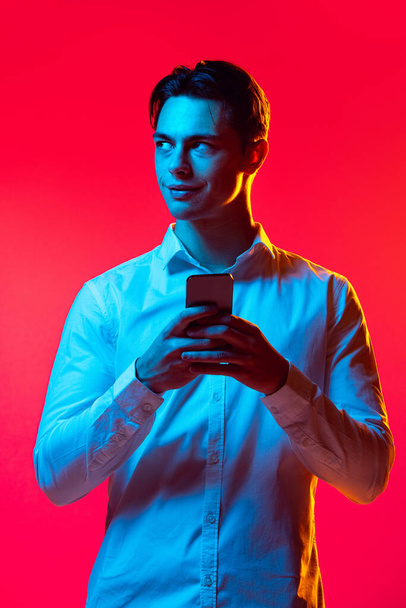 Portrait of young man in white shirt looks curious, typing phone message isolated over red studio background in blue neon - Foto, afbeelding