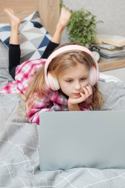 Uśmiechnięta dziewczynka korzystająca z laptopa w bezprzewodowych słuchawkach w łóżku w domu. - Zdjęcie, obraz