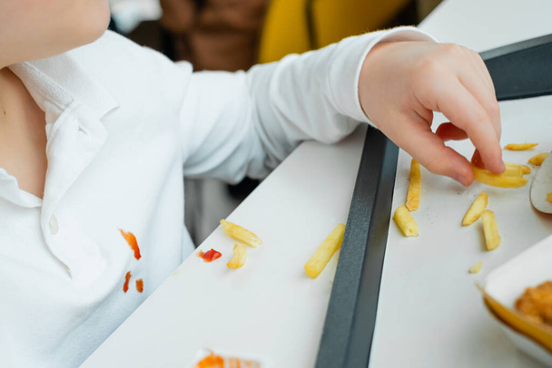 Children's hand holding french fries potato chips at the table in a fast food restaurant. Dirty stain of tomato sauce on clothes. High quality photo - Photo, Image