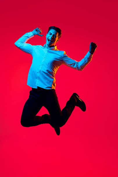 Portrait of young smiling man, student in white shirt cheerfully jumping in excitement isolated over red studio background in blue neon - Foto, imagen