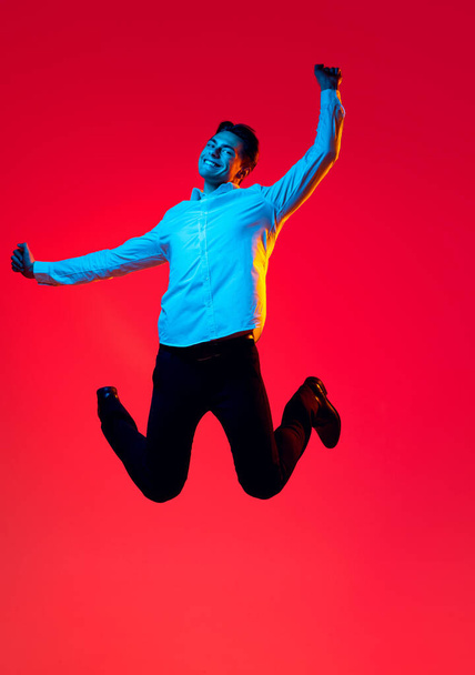 Portrait of young smiling man, student in white shirt cheerfully jumping in excitement isolated over red studio background in blue neon - Fotó, kép