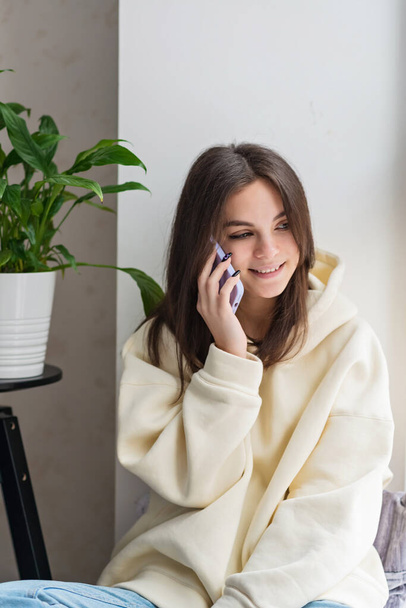Onnellinen iloinen nuori nainen puhuu puhelimessa kotona, hymyilee teini-ikäinen tyttö vastaa puheluun matkapuhelimeen. Pehmeä valikoiva kohdentaminen. - Valokuva, kuva