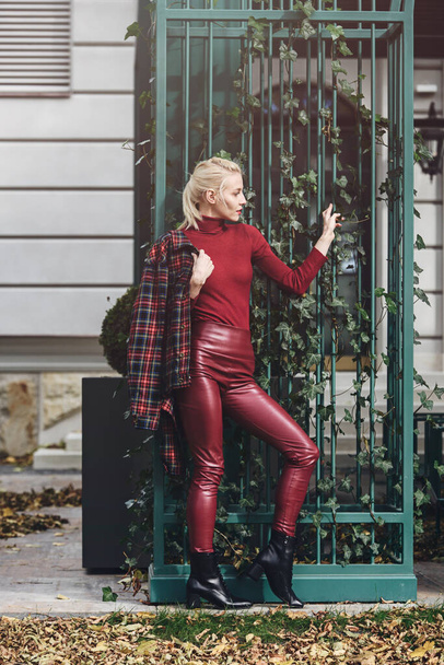 módní blondýnka s červenou rtěnkou pózující venku. Oblečený do červených kožených kamaší, roláků a kostkované bundy. sedící obrázek - Fotografie, Obrázek