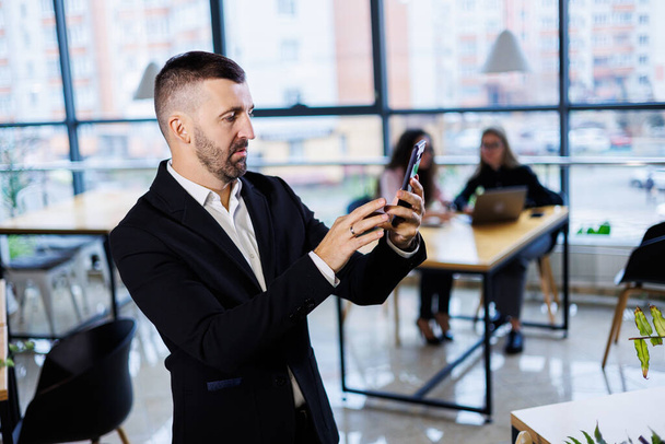 Портрет стильного молодого бізнесмена, що стоїть у великому сучасному офісі високо на верхньому поверсі і дивиться на телефон
 - Фото, зображення