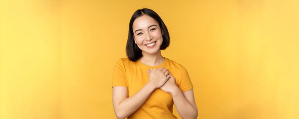 Romantyczna Koreanka, azjatka trzymająca się za serce, uśmiechnięta z troską i czułością, stojąca nad żółtym tłem - Zdjęcie, obraz