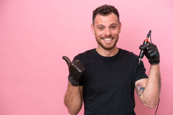 Tetování umělec brazilský muž izolované na růžovém pozadí ukazuje na stranu prezentovat produkt - Fotografie, Obrázek