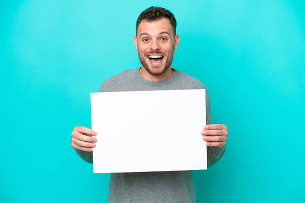 Elinde boş bir levha tutan genç Brezilyalı adam mavi arka planda izole edilmiş mutlu bir ifadeyle boş bir pankart tutuyor. - Fotoğraf, Görsel