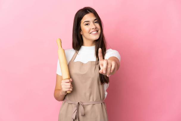 Teenager Brazilian girl holding a rolling pin isolated on pink background showing and lifting a finger - Photo, Image