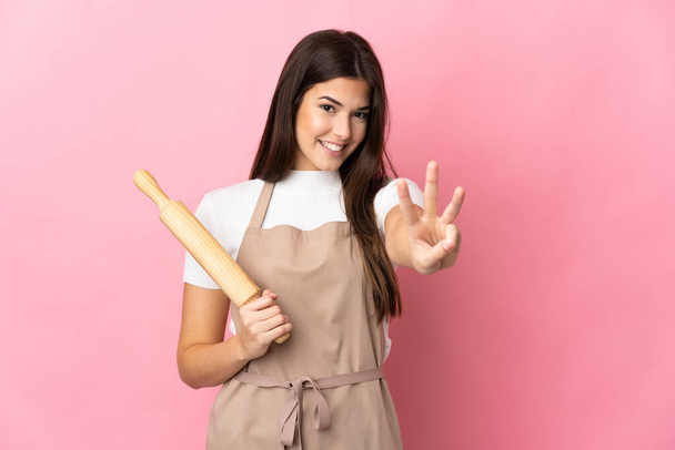 Teenager Brazilian girl holding a rolling pin isolated on pink background happy and counting three with fingers - Photo, Image