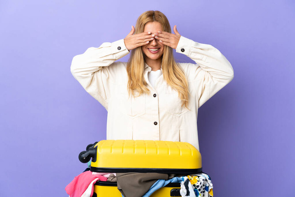 Młoda urugwajska blondynka z walizką pełną ubrań na odizolowanej fioletowej ścianie, okrywająca oczy dłońmi i uśmiechnięta - Zdjęcie, obraz