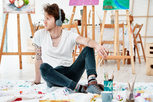 Inspiracja artystyczna poprzez muzykę. Pełne ujęcie przystojnego młodego artysty siedzącego samotnie i patrzącego kontemplacyjnie podczas słuchania muzyki przez słuchawki. - Zdjęcie, obraz