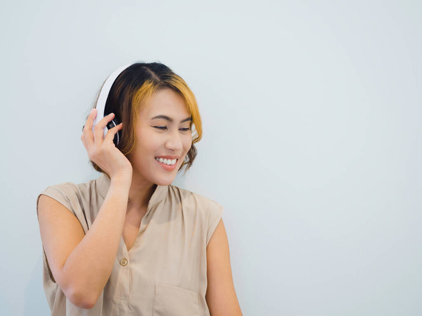 Onnellinen aasialainen nainen muotokuva lyhyet hiukset beige hihaton paita nauttia musiikin kuuntelusta, podcast, tai laulu radiossa valkoinen langaton kuuloke valkoisella taustalla kopioi tilaa. - Valokuva, kuva