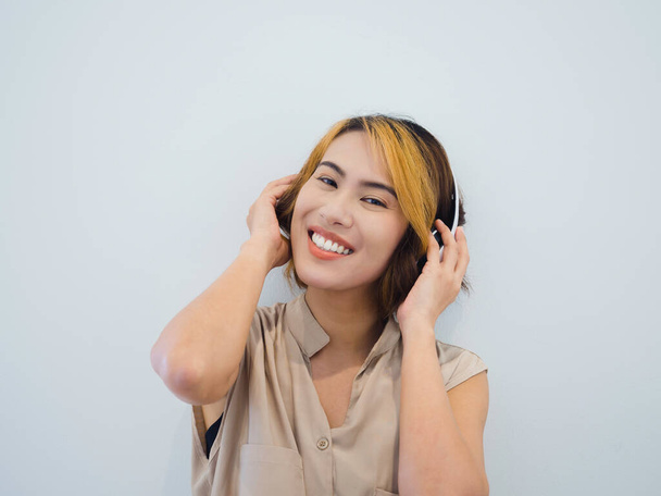 Happy Asian vrouw portret met kort haar in beige mouwloos shirt genieten van het luisteren naar muziek of lied op de radio met witte draadloze hoofdtelefoon op witte achtergrond, kijken naar camera. - Foto, afbeelding