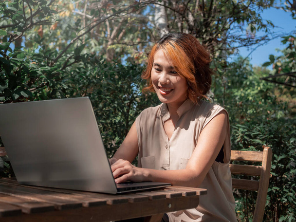 流行の短い髪の仕事と魅力的な幸せなアジアの女性の肖像画は、屋外の庭でリラックスした笑顔で木製の机の上に銀製のラップトップコンピュータ画面を見て。どこからでも. - 写真・画像