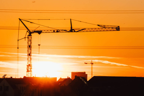 Lange bouwkraan silhouet in oranje hemel zonsondergang toont bouwplaats met engineering voor moderne gebouwen en stadsontwikkeling als architectonisch teamwork voor wolkenkrabbers hoogspanningslijnen - Foto, afbeelding