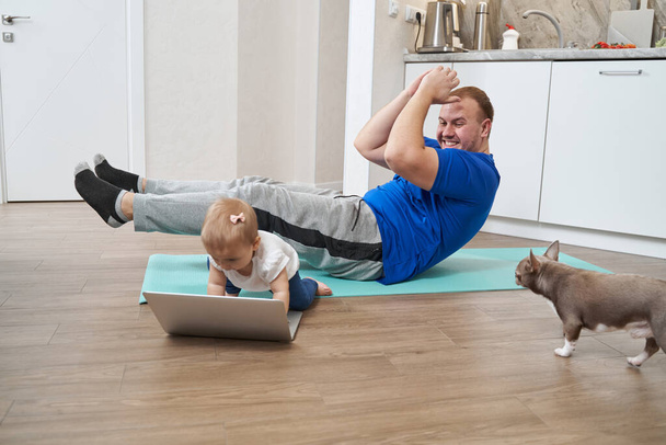 Homem fazendo crunches abdominais enquanto olha para o bebê perto do laptop - Foto, Imagem