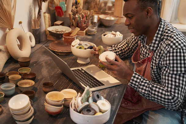 Joyous ceramist seated at laptop showing earthenware products to customer - Фото, зображення