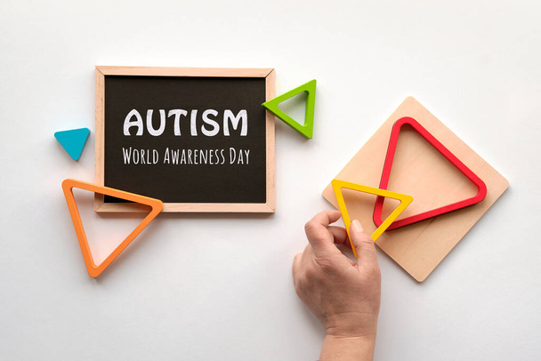 Text Autism awareness month on blackboard. Nested wood triangles in rainbow colors, human hand. Geometric education stacking puzzle. Montessori sensory toy. Flat lay on white paper background. - Foto, afbeelding