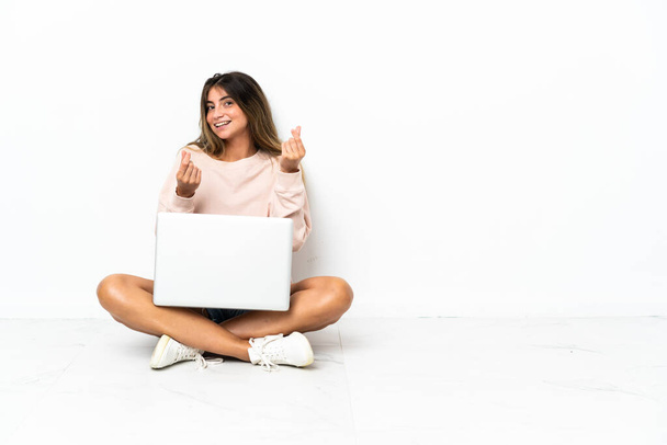 Молода жінка з ноутбуком, що сидить на підлозі ізольовано на білому тлі, заробляє жест грошей
 - Фото, зображення