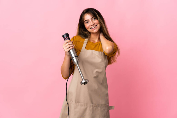 Joven chef mujer usando licuadora de mano aislada sobre fondo rosa riendo - Foto, Imagen