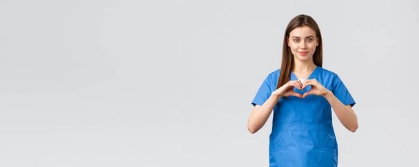 Trabajadores de la salud, prevenir virus, seguros y concepto de medicina. Una atractiva doctora sonriente, enfermera con uniformes azules se quedan a salvo en casa, muestran un signo cardíaco, expresan respeto a los combatientes del coronavirus
 - Foto, Imagen