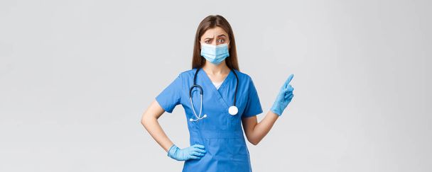 Covid-19, megelőzés vírus, egészségügyi dolgozók és karantén koncepció. Szkeptikus és elégedetlen női nővér vagy orvos kék köpenyben, orvosi maszk, mutogató ujj jobb, rosszalló kétkedő - Fotó, kép