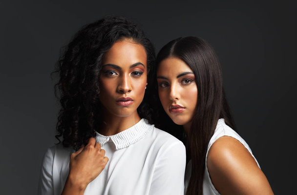 Women who stick together conquer together. Portrait of two attractive young women wearing white blouses posing closely together against a dark background in the studio. - Photo, Image