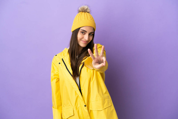 Mujer joven caucásica usando un abrigo impermeable aislado sobre fondo morado feliz y contando tres con los dedos - Foto, imagen