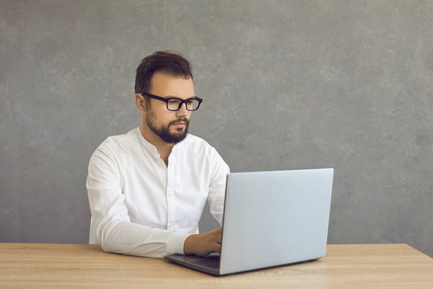 Young business man working on laptop sitting at desk against grey background - Valokuva, kuva