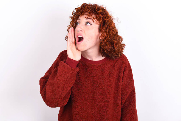 Молодая кавказская женщина с рыжими волосами в красном свитере на белом фоне продолжает кричать и громко кричать с поднятой рукой во рту. Концепция коммуникации. - Фото, изображение