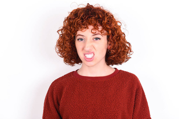 young caucasian woman red haired wearing red sweater over white background keeps keeps teeth clenched, frowns face in dissatisfaction, irritated because of much duties. - Photo, Image