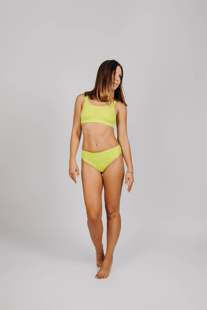 Woman poses in studio. Young brunette on light isolated background, stands in elegant pose on her toes, dressed in yellow underwear or swimsuit and demonstrates perfect slim, toned body - Fotó, kép